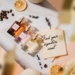 An overhead flat lay of three perfume bottles, dried citrus slices, coffee beans, lavender sprigs, and a note that reads 'Find your signature scent' on a marble and peach-colored background