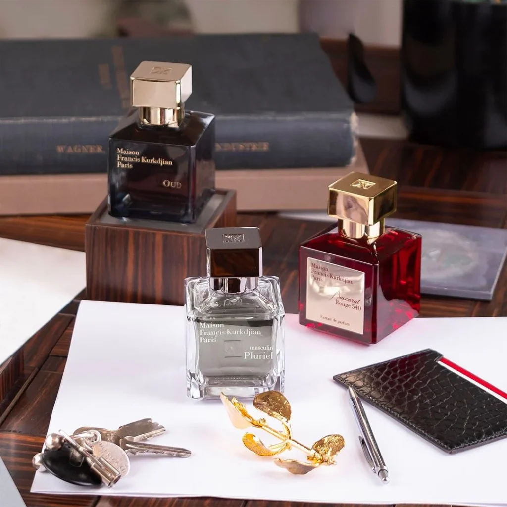 A stylish arrangement of three Maison Francis Kurkdjian niche perfume bottles—Oud, Masculin Pluriel, and Baccarat Rouge 540—placed on a wooden desk with accessories like keys, a notebook, and a gold leaf accent.