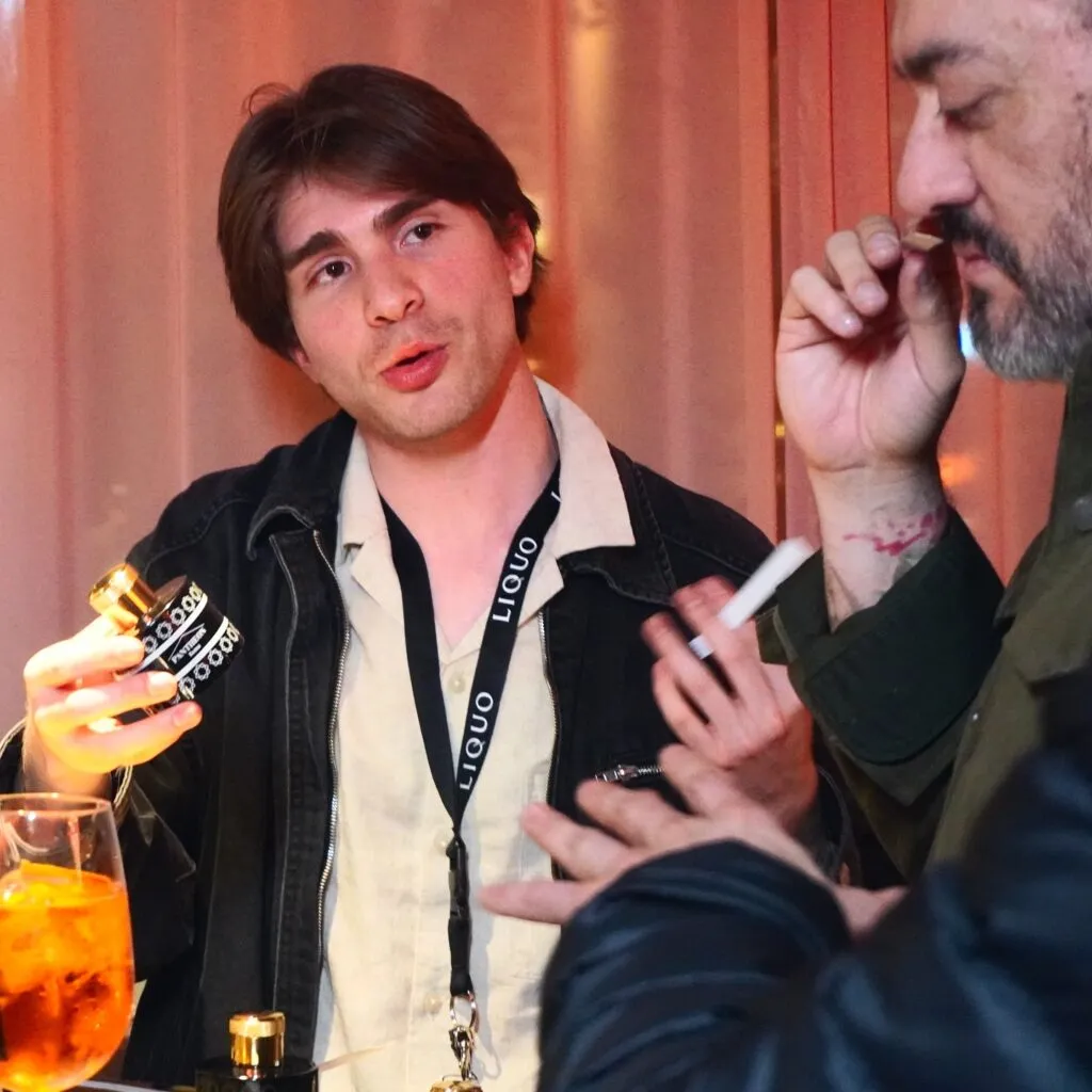 Presenting a perfume bottle at a LIQUO event, passionately discussing fragrance with a guest in an intimate, dimly lit setting.