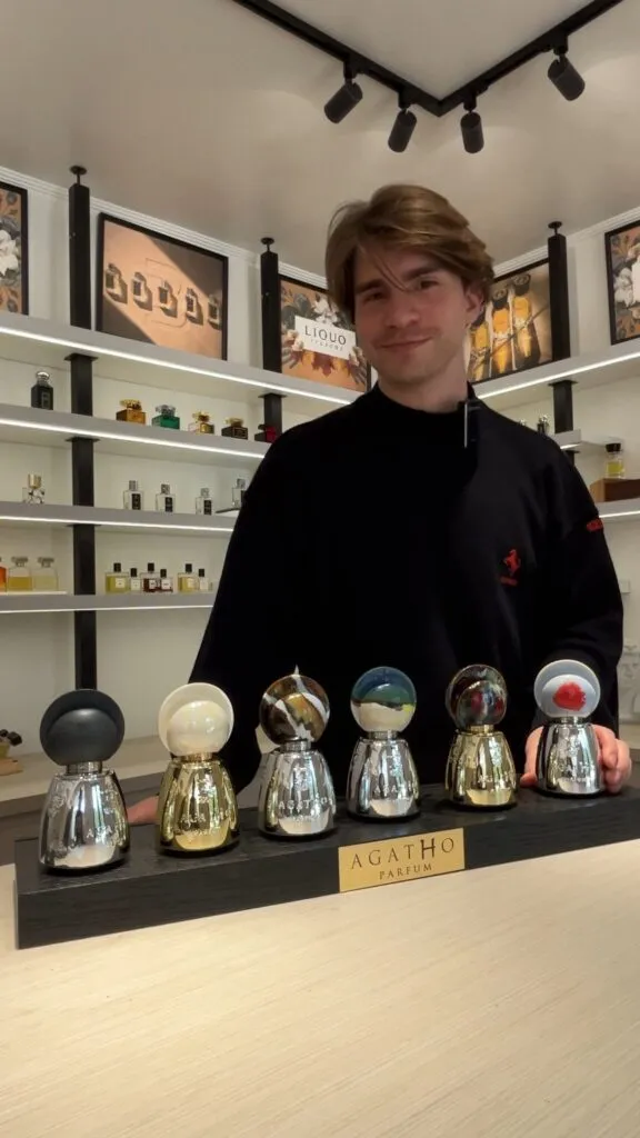 Standing behind a display of Agatho Parfum bottles at LIQUO, surrounded by shelves of niche fragrances, ready to share the art of perfumery and about me.
