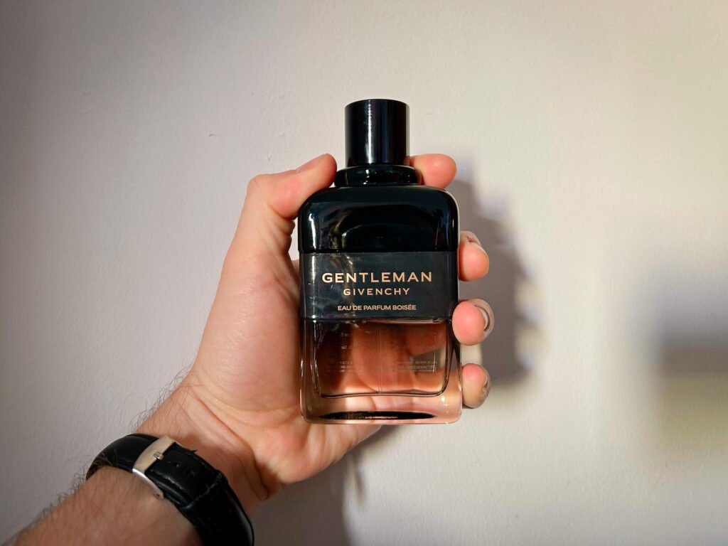 A hand holding a bottle of Givenchy Gentleman Eau de Parfum Boisée against a plain white wall. The black-to-brown gradient glass bottle has a sleek, elegant design with gold lettering on the label.