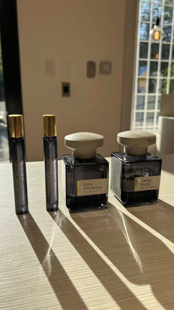 Sunlit tabletop display of four perfume vials—two Atelier Materi travel sprays and two square glass bottles labeled Bois d’Ambrette and Santal Blond Eau de Parfum.