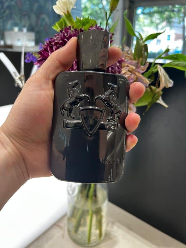 Hand holding a Herod by Parfums de Marly perfume bottle in natural light at Liquo, surrounded by flowers in the background.